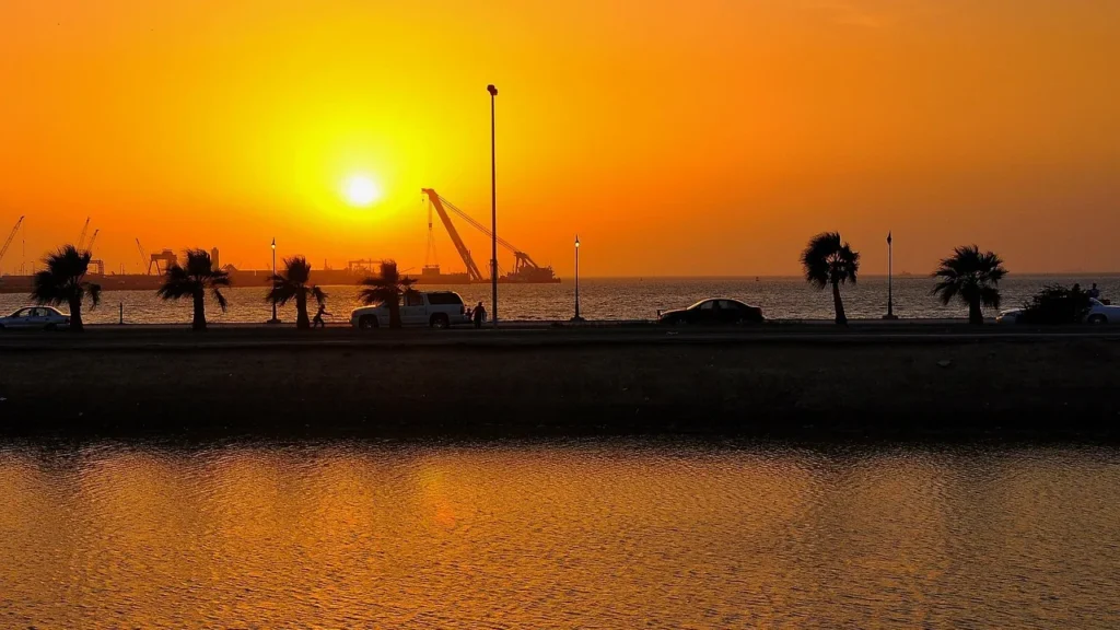 Puesta de sol anaranjada sobre el mar Rojo en la costa de Yeda, con palmeras y una carretera junto al mar a contraluz, una imagen de la mejor época para visitar Yeda