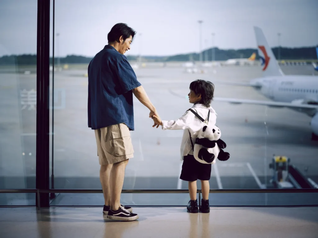 Un adulto y un niño de la mano junto a la ventana de un aeropuerto con aviones al fondo, antes de recoger a un menor no acompañado en Yeda