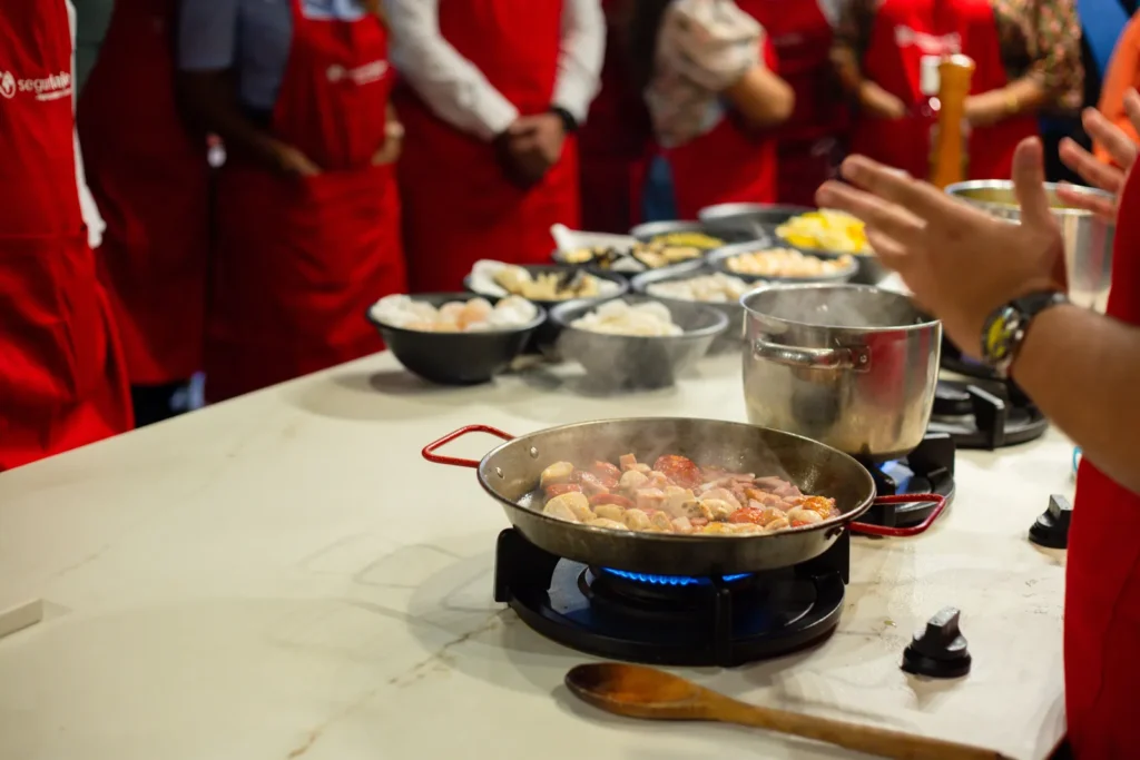 People in red aprons gathered around a counter with steaming cookware and a pan of seafood