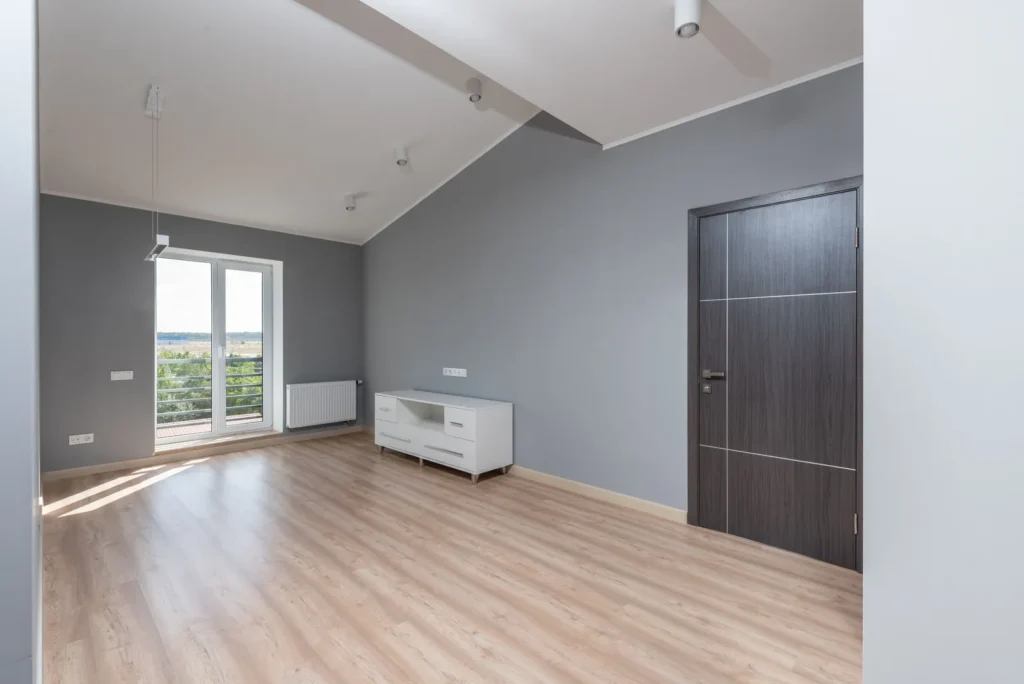 Una habitación casi vacía con suelo de madera, una puerta acristalada al balcón y un mueble bajo