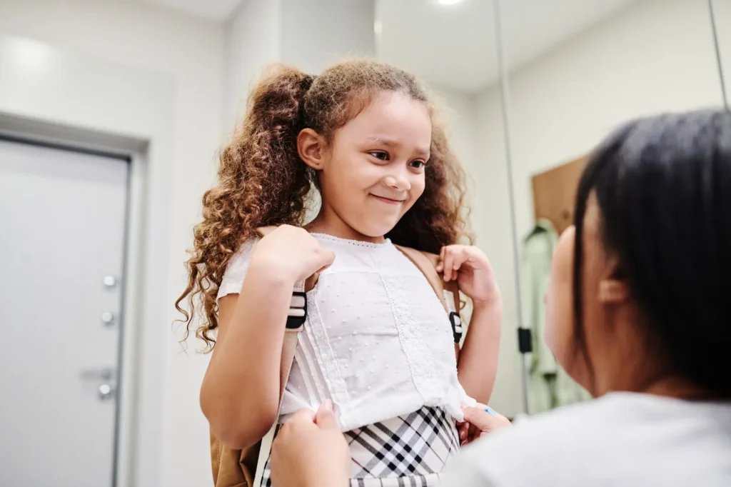 An adult helping a girl wearing a backpack indoors